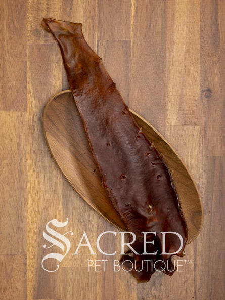 A long, narrow dried Aorta strip, a natural pet chew, laid diagonally on an oval wooden bowl over a wood plank surface. The Sacred Pet Boutique logo is printed at the bottom.