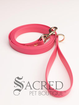 A fluro green biothane waterproof leash with brass hardware hanging from a brass hook on a white painted wooden post. It shows the full length of the leash which has it folded in half and the leash clip attached to the D ring at the leash handle. There is a beach in the background and a white Sacred Pet Boutique watermark the bottom.