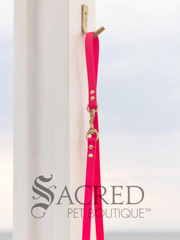 A fluro green biothane waterproof leash with brass hardware hanging from a brass hook on a white painted wooden post. It shows the full length of the leash which has it folded in half and the leash clip attached to the D ring at the leash handle. There is a beach in the background and a white Sacred Pet Boutique watermark the bottom.