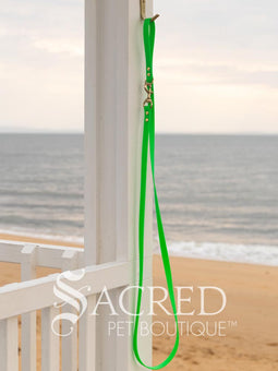 A fluro green biothane waterproof leash with brass hardware hanging from a brass hook on a white painted wooden post. It shows the full length of the leash which has it folded in half and the leash clip attached to the D ring at the leash handle. There is a beach in the background and a white Sacred Pet Boutique watermark the bottom.