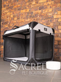 A Canine Care portable soft crate sits on a tile floor in a brick walled room. The side door has been rolled up. A white water bowl sits to the right of the crate. A white sherpa lining is inside the bottom of the crate. The crate looks spacious and airy.