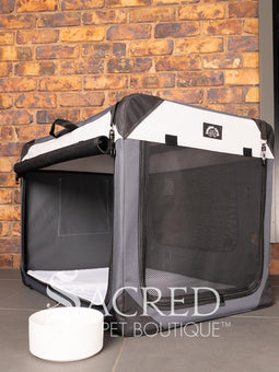 A Canine Care portable soft crate sits on a tile floor in a brick walled room. The side and an end door have been rolled up. An Italian Greyhound (it's Rossi) sits inside and looks to a person off screen. The crate looks spacious and airy.