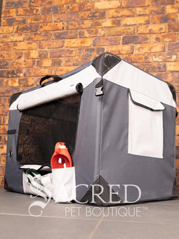 A Canine Care portable soft crate sits on a tile floor in a brick walled room. The side and an end door have been rolled up. An Italian Greyhound (it's Rossi) sits inside and looks to a person off screen. The crate looks spacious and airy.