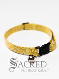 A cat collar is shown against a plain white background. The fabric has a white background, hardly noticeable, with naive yellow flowers overlapping each other. The collar has a black plastic breakaway buckle and adjuster, and a silver bell on a silver split ring.
