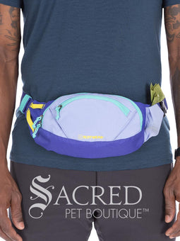 A person wearing a backpack with a blue and teal colour scheme is holding a treat in front of a dog's face. The dog is wearing a teal green harness and is attentive to the treat.