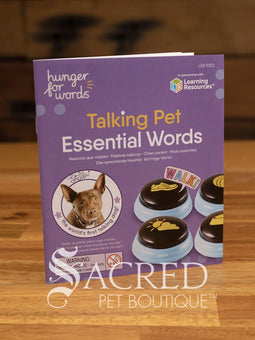 The product pack for Hunger for Words Talking Pet Essential Words sits on a wooden table with a wooden plank background. It shows the six pre-recorded blue and black buttons included, the words that are recorded, and pictures of dogs, although this product is suitable for cats.