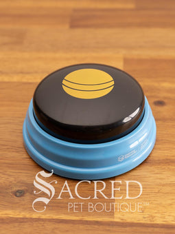 The product pack for Hunger for Words Talking Pet Essential Words sits on a wooden table with a wooden plank background. It shows the six pre-recorded blue and black buttons included, the words that are recorded, and pictures of dogs, although this product is suitable for cats.