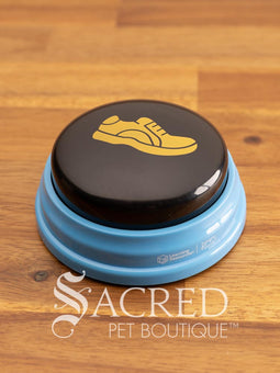 The product pack for Hunger for Words Talking Pet Essential Words sits on a wooden table with a wooden plank background. It shows the six pre-recorded blue and black buttons included, the words that are recorded, and pictures of dogs, although this product is suitable for cats.