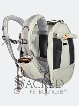Person carrying a pet carrier with a cat inside, with a dog. surrounded by greenery, featuring the watermark 'Sacred Pet Boutique'.
