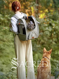 Person carrying a pet carrier with a cat inside, with a dog. surrounded by greenery, featuring the watermark 'Sacred Pet Boutique'.