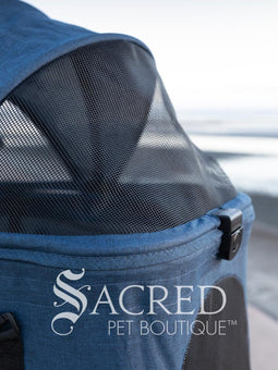 Pet stroller with a blue pet carrier on a paved path near a beach, featuring the Sacred Pet Boutique logo.