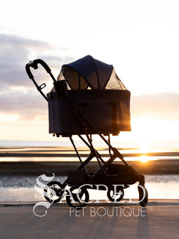 Pet stroller with a blue pet carrier on a paved path near a beach, featuring the Sacred Pet Boutique logo.