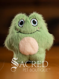 Three colorful plush toys (pink, yellow, green) on a wooden surface with 'Sacred Pet Boutique' branding.