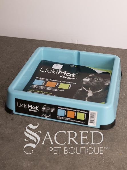 A light blue LickiMat Keeper pet lick mat tray with its retail packaging insert, placed on a gray countertop against a white wall. A watermark for 'Sacred Pet Boutique' is visible at the bottom.