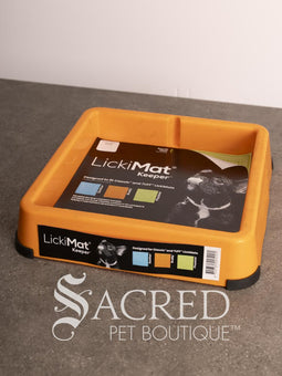 A light blue LickiMat Keeper pet lick mat tray with its retail packaging insert, placed on a gray countertop against a white wall. A watermark for 'Sacred Pet Boutique' is visible at the bottom.