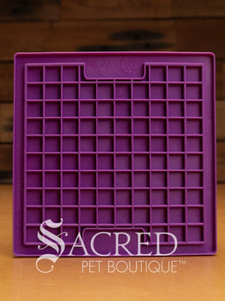 A purple LickiMat Boredom Buster designed for pets, with a textured surface designed to promote licking and calm behaviour. This design is the Classic Buddy and features a distinct criss cross matrix pattern designed to hold soft spreadable treats. It is on a wooden surface with a dark wood background and has a white Sacred Pet Boutique watermark.