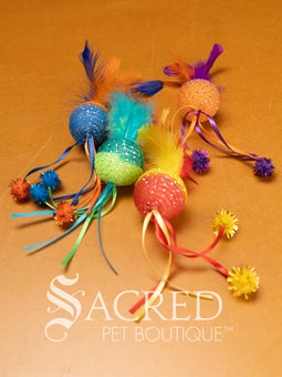 A selection of colourful mesh ball cat toys with ribbons and feather on a wooden background with a white Sacred Pet Boutique watermark.