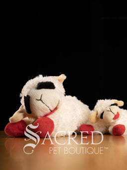 Two plush Lamb Chop sheep dog toys with red feet and tongues on a wooden surface, branded 'Sacred Pet Boutique'.