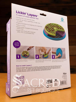 A licking layers toy is shown on a wooden table with a wooden plank wall background. You can clearly see the three different layers of the toy. The top layer is a green hexagonal design. The middle layer is a purple circle design. The bottom layer is blue square design.