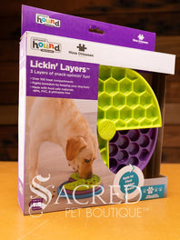 A licking layers toy is shown on a wooden table with a wooden plank wall background. You can clearly see the three different layers of the toy. The top layer is a green hexagonal design. The middle layer is a purple circle design. The bottom layer is blue square design.