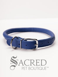 A black, round, soft leather dog collar with a metal buckle clasp. Our beautiful Soft Round (Rolled) leather collars are highly recommended for long haired dogs. Round or rolled collars reduce the amount of matting, tangling and flattening of the dogs fur. All collars are hand made and stitched with wax thread, from the finest quality soft leather wrapped and stitched around a nylon inner cord for added strength.Image on a white background, with the Sacred Pet Boutique logo.