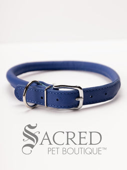 A black, round, soft leather dog collar with a metal buckle clasp. Our beautiful Soft Round (Rolled) leather collars are highly recommended for long haired dogs. Round or rolled collars reduce the amount of matting, tangling and flattening of the dogs fur. All collars are hand made and stitched with wax thread, from the finest quality soft leather wrapped and stitched around a nylon inner cord for added strength.Image on a white background, with the Sacred Pet Boutique logo.