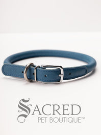 A black, round, soft leather dog collar with a metal buckle clasp. Our beautiful Soft Round (Rolled) leather collars are highly recommended for long haired dogs. Round or rolled collars reduce the amount of matting, tangling and flattening of the dogs fur. All collars are hand made and stitched with wax thread, from the finest quality soft leather wrapped and stitched around a nylon inner cord for added strength.Image on a white background, with the Sacred Pet Boutique logo.