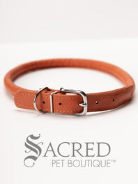 A black, round, soft leather dog collar with a metal buckle clasp. Our beautiful Soft Round (Rolled) leather collars are highly recommended for long haired dogs. Round or rolled collars reduce the amount of matting, tangling and flattening of the dogs fur. All collars are hand made and stitched with wax thread, from the finest quality soft leather wrapped and stitched around a nylon inner cord for added strength.Image on a white background, with the Sacred Pet Boutique logo.