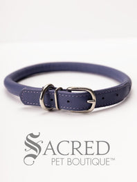 A black, round, soft leather dog collar with a metal buckle clasp. Our beautiful Soft Round (Rolled) leather collars are highly recommended for long haired dogs. Round or rolled collars reduce the amount of matting, tangling and flattening of the dogs fur. All collars are hand made and stitched with wax thread, from the finest quality soft leather wrapped and stitched around a nylon inner cord for added strength.Image on a white background, with the Sacred Pet Boutique logo.