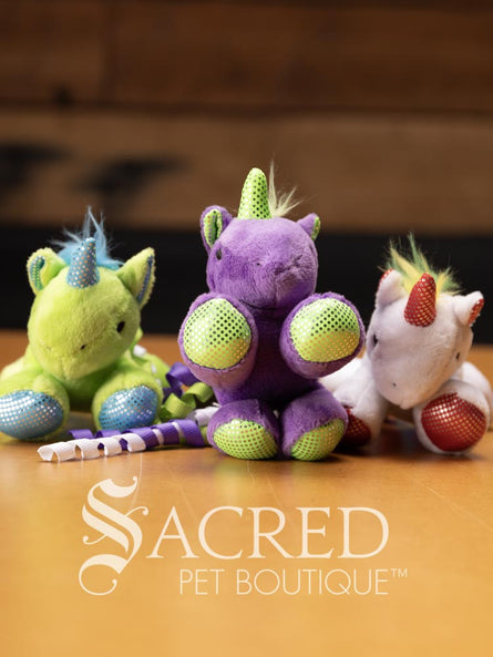 Three plush unicorn cat toys in different colours, each with a ribbon tail and glittery spots, placed on a wooden surface with a 'Sacred Pet Boutique' watermark.