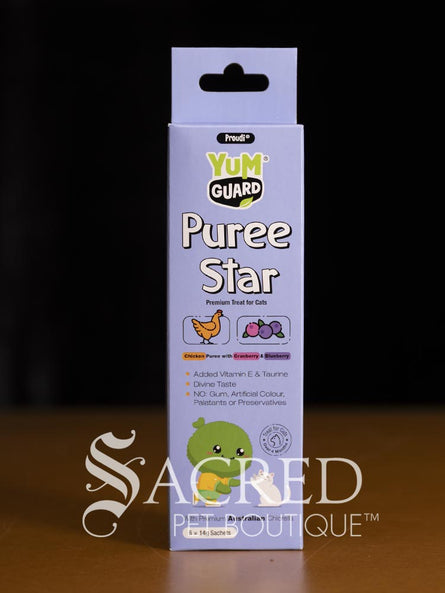 A boxed pet product labeled Yumguard Puree Star stands upright on a wooden surface against a dark background. The package is predominantly white and light purple/lavender with green branding and small food icons. With a white Sacred Pet Boutique watermark.