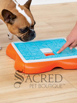An orange puzzle feeder with blue tiles that slide along tracks with a white plastic bone shape in the centre. Sliding out from this is a bright orange section that has circular compartments meant for holding treats. On the right hand side of the image a person's hand is visible placing small, brown treats treats in to the empty compartments. This toy is lying on a pale wood floor and a grey Sacred Pet Boutique watermark is at the bottom.