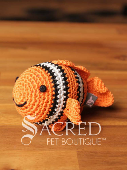 A cute crocheted orange, black and white clown fish rests on a wooden table with a wooden wall behind. Suitable for small, toy and teacup dogs, the crochet toys squeak and will floss teeth as they play.