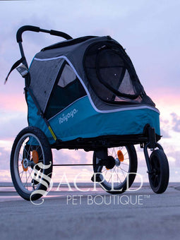 The Happy Pet 3 wheeled bicycle trailer and stroller, by Ibiyaya is a dual-purpose pet stroller that can attach to your bike. Shown in Ocean Blue colour includes bicycle attachment and rain cover. Photo of stroller on ground with cloudy sky in background with Sacred Pet Boutique watermark.