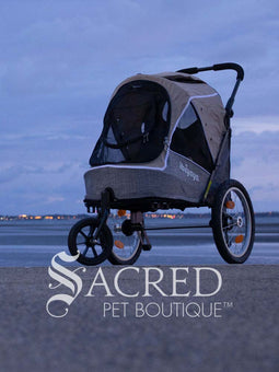 The Happy Pet 3 wheeled bicycle trailer and stroller, by Ibiyaya is a dual-purpose pet stroller that can attach to your bike. Shown in Ocean Blue colour includes bicycle attachment and rain cover. Photo of stroller on ground with cloudy sky in background with Sacred Pet Boutique watermark.