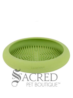 A white dog is shown in a stainless steel laundry sink being washed and he is licking a blue Lickimat UFO bowl that is stuck to the white tiled wall in front of him. This is being used to distract him whilst he is being groomed. At the bottom on of the image is a grey Sacred Pet Boutique watermark.
