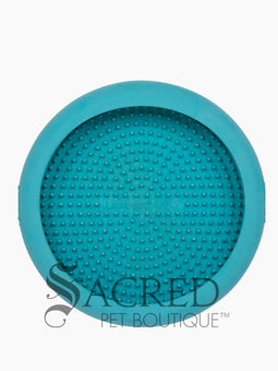 A white dog is shown in a stainless steel laundry sink being washed and he is licking a blue Lickimat UFO bowl that is stuck to the white tiled wall in front of him. This is being used to distract him whilst he is being groomed. At the bottom on of the image is a grey Sacred Pet Boutique watermark.