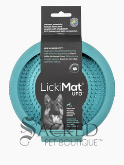 A white dog is shown in a stainless steel laundry sink being washed and he is licking a blue Lickimat UFO bowl that is stuck to the white tiled wall in front of him. This is being used to distract him whilst he is being groomed. At the bottom on of the image is a grey Sacred Pet Boutique watermark.