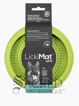A white dog is shown in a stainless steel laundry sink being washed and he is licking a blue Lickimat UFO bowl that is stuck to the white tiled wall in front of him. This is being used to distract him whilst he is being groomed. At the bottom on of the image is a grey Sacred Pet Boutique watermark.