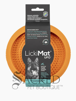 A white dog is shown in a stainless steel laundry sink being washed and he is licking a blue Lickimat UFO bowl that is stuck to the white tiled wall in front of him. This is being used to distract him whilst he is being groomed. At the bottom on of the image is a grey Sacred Pet Boutique watermark.