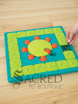 A teal, lime green and orange plastic interactive dog puzzle toy placed on a light coloured wooden floor. The toy is square shaped with a teal base. It has an outer track which is lined with square lime green tiles that slide to reveal hidden treats. There is a central lime green wheel that has a curved indentation and is surrounded by orange pegs. To the right there is a hand dropping treats into a space that can be hidden. At the bottom of the image is a grey Sacred Pet Boutique watermark.