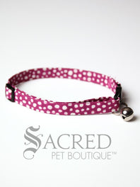 A cat collar is shown against a plain white background. The pattern of the fabric is pink with white polker dots. The collar has a black plastic breakaway buckle and adjuster, and a silver bell on a silver split ring.