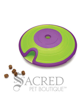 A green and purple saucer shaped interactive dog toy. It features a lime green outer ring and a vibrant purple centre and rim. A specialised curved indentation or track is visible on the green sections which allows treats to fall out. Near this opening is five dog treats showing how they fall out. At the bottom of this image is a grey Sacred Pet Boutique watermark.