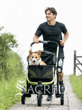 A man jogging on a paved path with a corgi in a black pet stroller with lime green accents. Includes a drink/bottle holder. A watermark for 'Sacred Pet Boutique' is visible at the bottom.