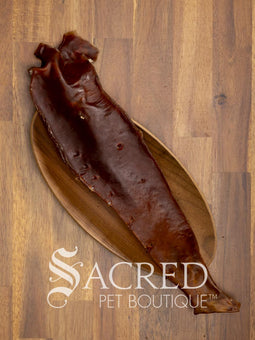 A long, narrow dried Aorta strip, a natural pet chew, laid diagonally on an oval wooden bowl over a wood plank surface. The Sacred Pet Boutique logo is printed at the bottom.