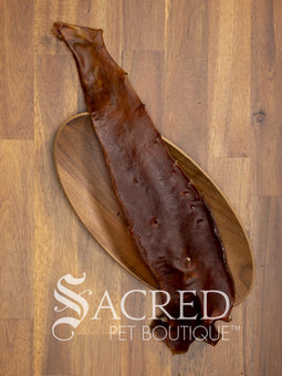 A long, narrow dried Aorta strip, a natural pet chew, laid diagonally on an oval wooden bowl over a wood plank surface. The Sacred Pet Boutique logo is printed at the bottom.