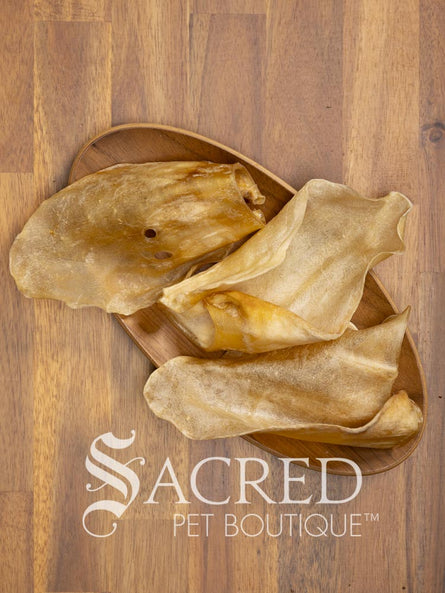 Several dried, translucent beef ear dog treats are arranged on a small oval wooden plate over a wood-grain surface. The brand name 'Sacred Pet Boutique' is printed in white at the bottom.