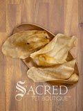 Several dried, translucent beef ear dog treats are arranged on a small oval wooden plate over a wood-grain surface. The brand name 'Sacred Pet Boutique' is printed in white at the bottom.
