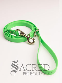 A fluro green biothane waterproof leash with brass hardware hanging from a brass hook on a white painted wooden post. It shows the full length of the leash which has it folded in half and the leash clip attached to the D ring at the leash handle. There is a beach in the background and a white Sacred Pet Boutique watermark the bottom. 