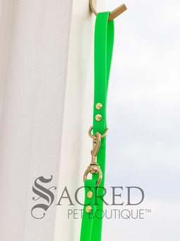 A fluro green biothane waterproof leash with brass hardware hanging from a brass hook on a white painted wooden post. It shows the full length of the leash which has it folded in half and the leash clip attached to the D ring at the leash handle. There is a beach in the background and a white Sacred Pet Boutique watermark the bottom. 